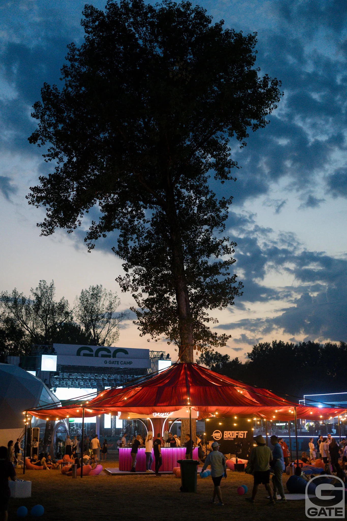 The outdoor business festival at dusk beneath a large tree, with G GATE CAMP structures in the background.
